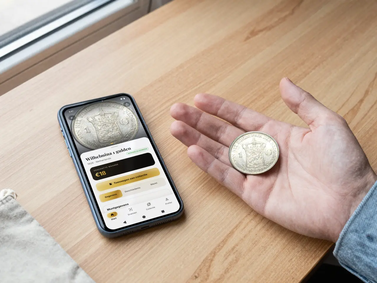 An open hand holding a silver 1929 guilder next to a phone on the table. The screen shows Wilhelmina 1 gulden, 1929, uncommon, estimated value €18.
